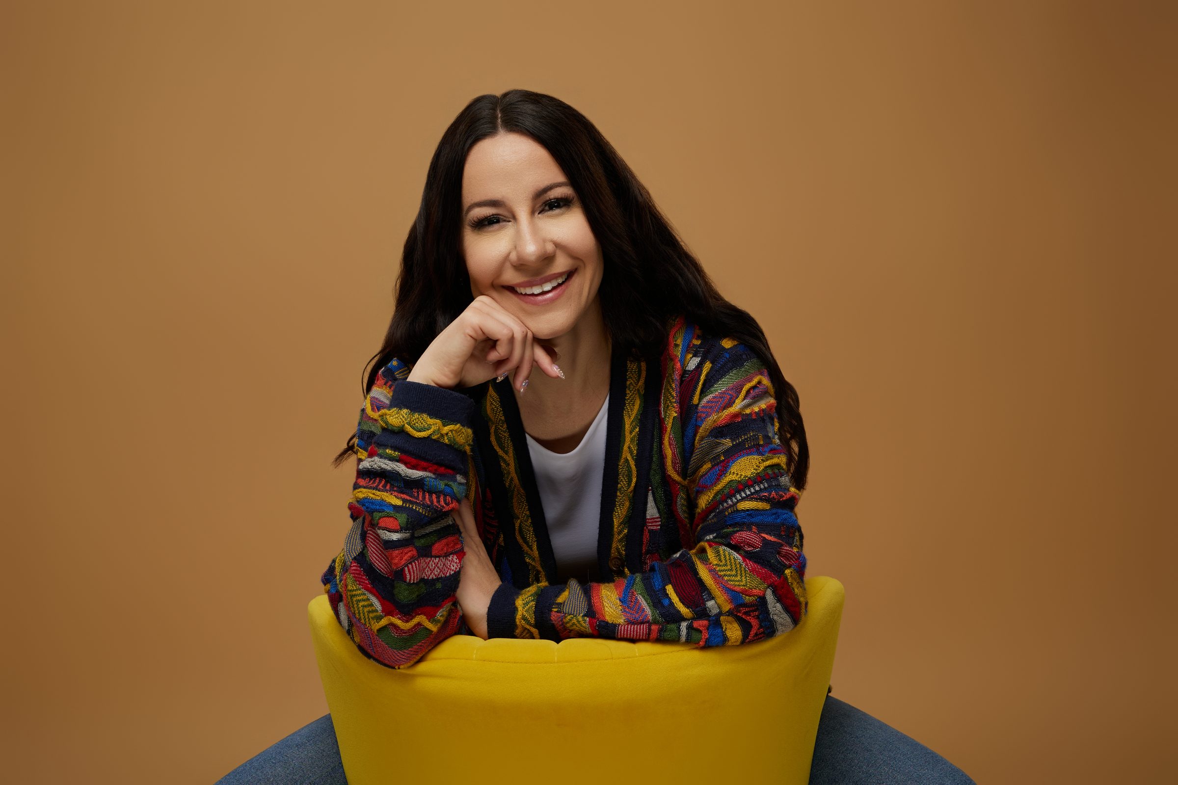 Bryanna Baptiste in her Coogi cardigan, seated in a yellow chair with her chin resting on her hand, against a caramel backdrop